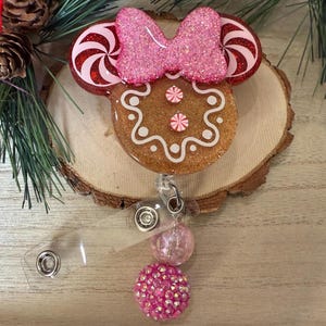 May include: A gingerbread-themed Minnie Mouse-shaped badge holder with a pink bow, candy cane ears, and pink and white glitter. The holder is attached to a clear retractable badge reel with two pink and white glitter charms.