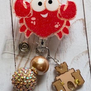 May include: A red glitter crab badge reel with a smiling face and white eyes. The badge reel has a charm of a sandcastle, a pearl, and a decorative bead. The badge reel is attached to a clear plastic reel.