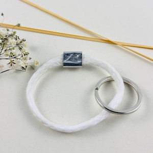 Pode incluir: Chaveiro de corda branca com um anel prateado e um charme quadrado prateado. O charme tem um design texturizado.