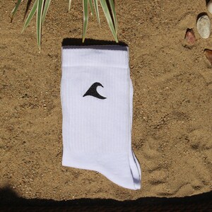 Könnte beinhalten: Weiße Socken mit einem schwarzen Wellenlogo auf der Vorderseite. Die Socken liegen auf einer sandigen Oberfläche.