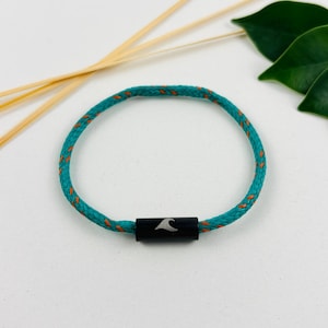 Op de afbeelding: Turquoise touw armband met oranje stiksels en een zwarte sluiting met een wit golfontwerp. De armband is cirkelvormig en ligt op een wit oppervlak, met decoratieve bamboestokken en groene bladeren op de achtergrond.