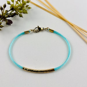 Könnte beinhalten: Ein türkisfarbenes geflochtenes Armband aus Kordel mit einem goldenen, gehämmerten Balkenakzent. Das Armband hat einen Karabinerverschluss.