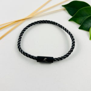 Könnte beinhalten: Ein schwarz-graues geflochtenes Armband mit einem Wellenmuster auf dem Verschluss. Das Armband besteht aus einem seilartigen Material und ist kreisförmig. Der Hintergrund umfasst grüne Blätter und dünne, hellfarbene Stäbe.