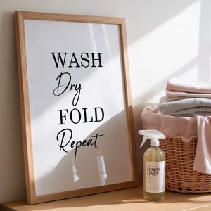 Könnte beinhalten: Ein gerahmter Druck mit den Worten "WASH Dry FOLD Repeat" in schwarzer Schrift. Eine Flasche Lemon Linen Spray und ein Weidenkorb mit gefalteter Wäsche stehen auf einer Holzoberfläche.