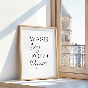 May include: A rectangular wall art print in a light wood frame featuring the black text WASH, Dry, FOLD, Repeat centered on a white background. The print is displayed upright, showcasing a clean design with a mix of serif and script fonts.