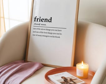 May include: A framed print with the definition of "friend" in black text. The print is in a light wood frame and rests on a white chair. A lit candle and a book are on a pink tray.