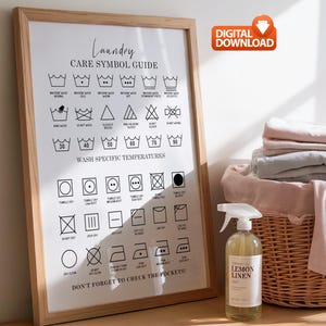 May include: A framed laundry care symbol guide with a wooden frame. The guide includes various washing instructions and temperature settings. A bottle of "Lemon Linen" spray sits next to the frame. A basket of folded linens is in the background.