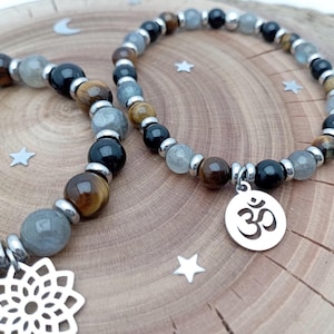 Op de afbeelding: Twee armbanden met kralen in zwart, grijs, bruin en tijgeroog. Eén armband heeft een zilveren bedel met het Om-symbool, de andere een bloembedel. De armbanden liggen op een houten ondergrond.