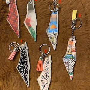 Könnte beinhalten: Sechs Schlüsselanhänger mit verschiedenen Designs des palästinensischen Gebiets. Jeder Schlüsselanhänger hat einen andersfarbigen Quasten und einen silbernen Ring. Die Designs umfassen ein geometrisches Muster, eine palästinensische Flagge, eine Moschee, eine Stadtlandschaft, arabische Kalligraphie und eine Kombination aus arabischer Kalligraphie und einer Moschee.