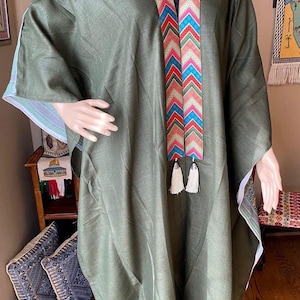 May include: Olive green caftan with a colorful chevron pattern along the neckline and down the front. The caftan has wide sleeves and white trim. Two white tassels hang from the bottom of the chevron design. The caftan is displayed on a mannequin.