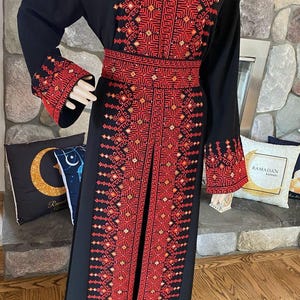 May include: A long black abaya with a red and orange embroidered design. The abaya has a high neckline and long sleeves. The embroidery is intricate and detailed, and it covers the entire front of the abaya.