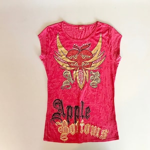 Può includere: Una maglietta rosa a maniche corte con un design grafico oro e rosso. Il design presenta una mela con le ali e le lettere "AMB". Le parole "Apple Bottoms" sono stampate in oro e nero.