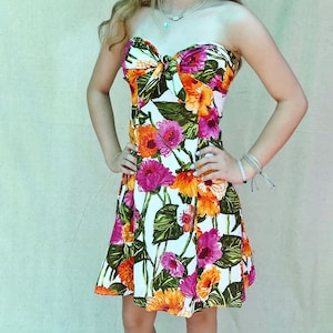 May include: A strapless dress with a vibrant floral print featuring pink, orange, and yellow flowers on a white background. The dress has a fitted bodice with a bow detail and a flared skirt. Green leaves are interspersed among the flowers.