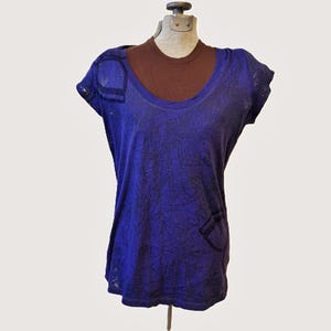 May include: A layered top on a mannequin. The outer layer is a short-sleeved, deep blue t-shirt with a scoop neck and a subtle, textured pattern. A brown undershirt is visible beneath. The t-shirt has a pocket design.