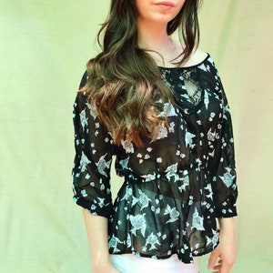 May include: Black sheer blouse with a floral pattern in white and gray. The blouse has three-quarter sleeves, a round neckline, and a cinched waist. The top is made of a lightweight fabric.