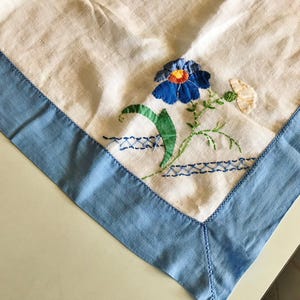 Puede incluir: Un mantel de lino blanco con un borde azul claro y detalles florales bordados. El bordado presenta una flor azul con un centro naranja, hojas verdes y una pequeña flor blanca. El borde es de color azul claro liso.