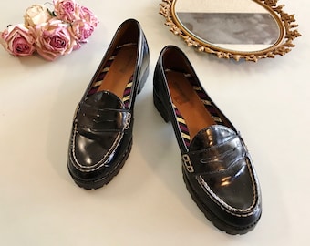 May include: Pair of black leather penny loafers with white stitching and a chunky sole. The shoes have a glossy finish and a striped interior. A classic style of footwear.