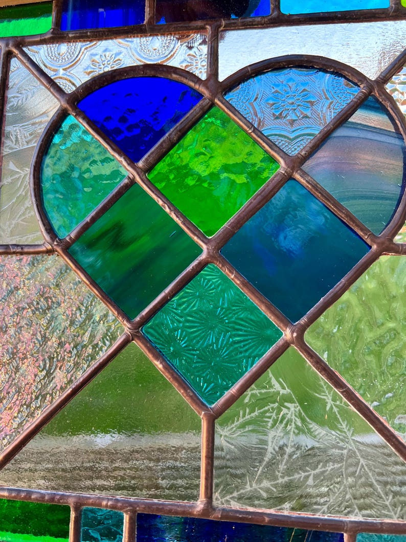 May include: A stained glass window with a heart-shaped design. The heart is made up of blue, green, and clear glass pieces. The glass is textured and has a vintage look.