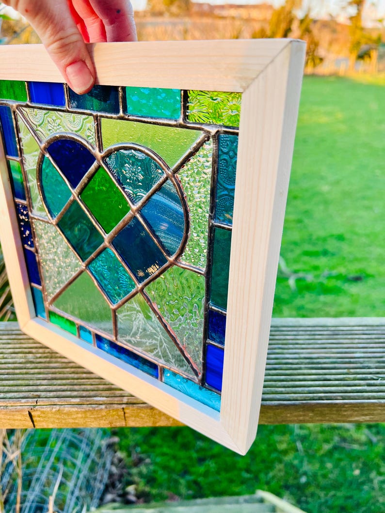 May include: A stained glass window panel with a heart shape design in shades of blue and green. The panel is framed in natural wood.
