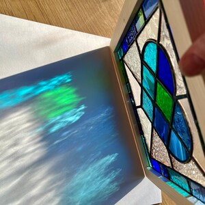 May include: A stained glass window with a heart design in shades of blue, green, and white. The window is framed in wood and casts a colorful shadow on a white surface.