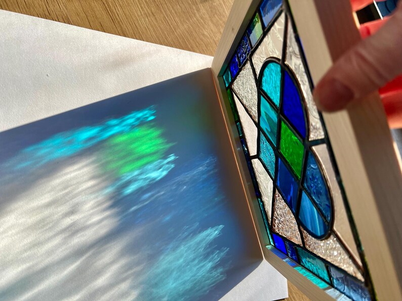 May include: A stained glass window with a heart design in shades of blue, green, and white. The window is framed in wood and casts a colorful shadow on a white surface.
