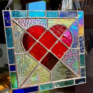 May include: A square stained glass panel with a red heart at its centre. The heart is surrounded by colourful glass pieces in shades of blue, green, yellow, and iridescent hues. The panel is framed with silver-coloured metal and has a black hanging ribbon.