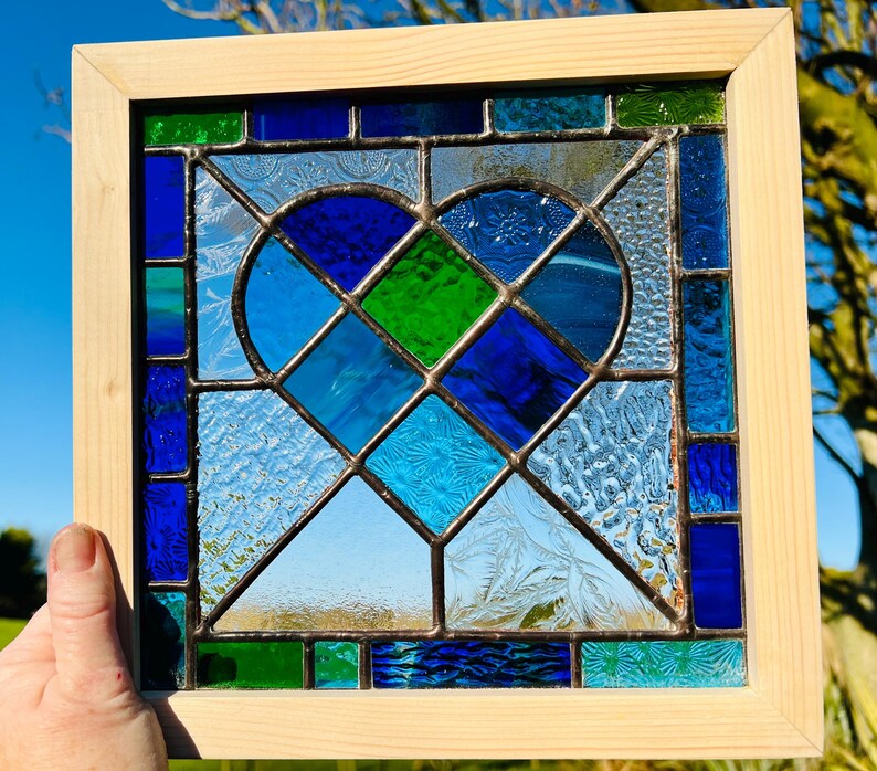 May include: A stained glass window panel featuring a blue and green heart design. The heart is made up of multiple pieces of glass, creating a geometric pattern. The panel is framed in wood.
