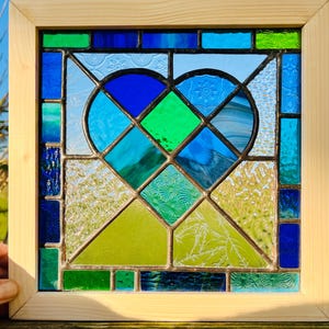 May include: A stained glass window panel featuring a heart shape made of blue, green, and white glass pieces. The heart is surrounded by a frame of wood.