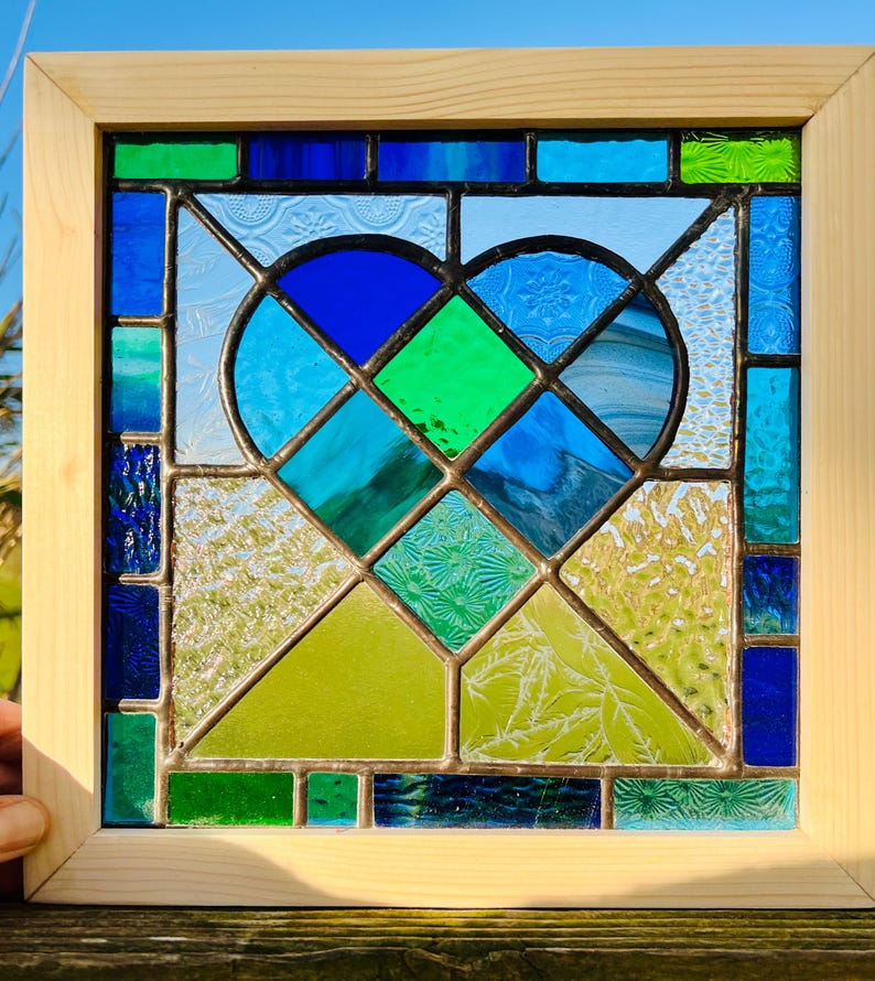 May include: A stained glass window panel featuring a heart shape made of blue, green, and white glass pieces. The heart is surrounded by a frame of wood.