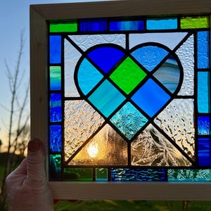 May include: A stained glass window panel featuring a blue and green heart design. The heart is made up of smaller pieces of glass in various shades of blue and green, with a textured glass background.