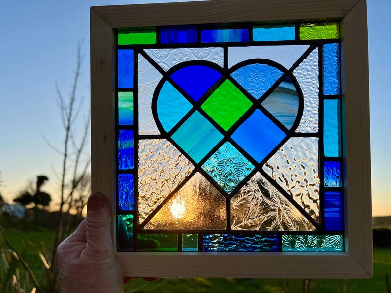 May include: A stained glass window panel featuring a blue and green heart design. The heart is made up of smaller pieces of glass in various shades of blue and green, with a textured glass background.