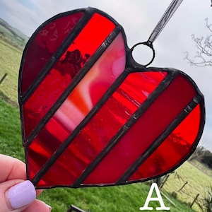 May include: A red and pink stained glass heart with a striped pattern. The heart is edged in black and has a black ribbon for hanging. The letter "A" is visible in the lower right corner.