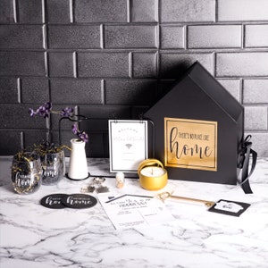 May include: A housewarming gift box with a black and gold theme. The box is shaped like a house and has a gold sticker that says "There's no place like home". The box contains a candle, a key, coasters, and other housewarming gifts.