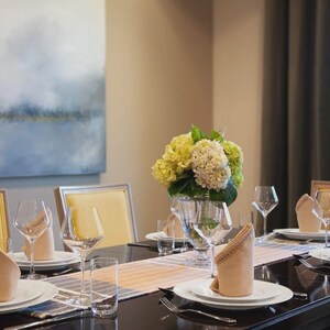 May include: Elegant dining table setting with white plates, wine glasses, and folded napkins. A centerpiece of yellow and green flowers sits in a clear vase. The table is set for a formal meal.