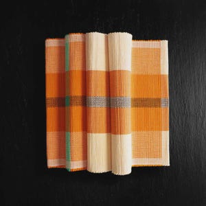 May include: Rolled orange and cream-colored placemats with a woven texture. The placemats have a striped design with accents of green and gray. The placemats are displayed on a black surface.