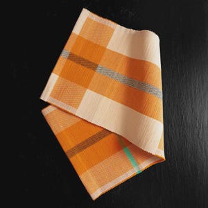 May include: Two folded orange and cream-colored woven placemats with a plaid pattern. The placemats have a textured appearance and feature thin stripes of gray and teal. The background is a dark, solid color.