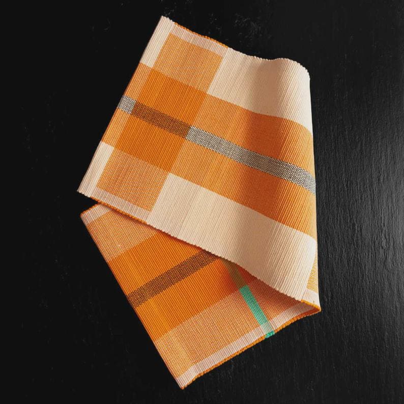 May include: Two folded orange and cream-colored woven placemats with a plaid pattern. The placemats have a textured appearance and feature thin stripes of gray and teal. The background is a dark, solid color.