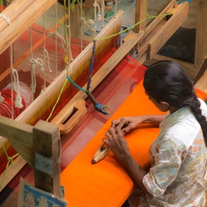 May include: A person weaving on a wooden loom. The loom has red, orange, and yellow threads. The person is wearing a light-colored floral shirt and has long dark hair. The woven fabric is a vibrant orange color.