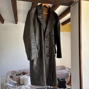 May include: A long, dark brown leather coat with a double-breasted closure and a collar. The coat has a worn, vintage look.