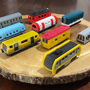 Puede incluir: Una colección de nueve vagones de tren de juguete en miniatura de varios colores, incluyendo gris, azul, rojo, blanco, amarillo y azul verdoso. Los vagones están dispuestos sobre una superficie de madera.