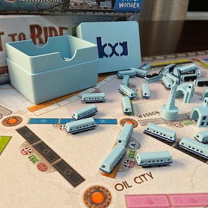 May include: A light blue board game set with train cars, buildings, and a card holder. The game board features a map with city names. The set includes a box with the letters "bca" on it. The game is "Ticket to Ride".