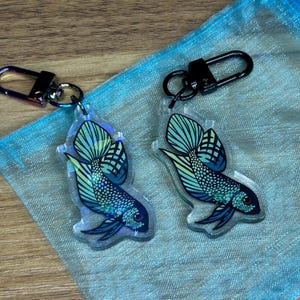 Könnte beinhalten: Zwei schillernde blau-grüne Betta-Fisch-Schlüsselanhänger mit schwarzen Metallschlüsselringen. Die Schlüsselanhänger befinden sich auf einem blauen und weißen Netzbeutel.