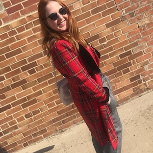 Puede incluir: Una mujer con un abrigo rojo y negro a cuadros, pantalones grises y zapatos azul oscuro. Está de pie frente a una pared de ladrillo.