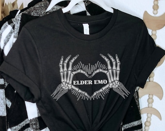 May include: Black t-shirt featuring a heart made of skeleton hands with the text "ELDER EMO" in the center. The shirt is hanging on a white hanger with a black and white plaid jacket in the background.