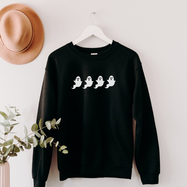 Op de afbeelding: Zwart sweatshirt met een wit spookontwerp. Het ontwerp toont vier witte spookfiguren op een rij. Het sweatshirt hangt aan een witte hanger tegen een witte muur. Een bruine hoed is zichtbaar op de achtergrond.