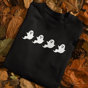 Op de afbeelding: Zwart sweatshirt met een ontwerp van vier witte spookfiguren. Elke geest heeft een eenvoudige, afgeronde vorm met ovale ogen en open mond. Het kledingstuk wordt getoond op een bed van herfstbladeren en dennenappels.