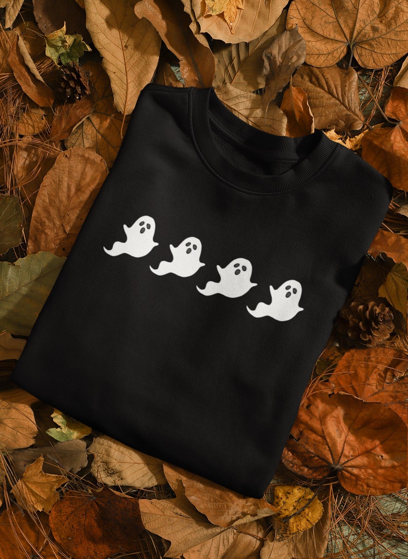 Op de afbeelding: Zwart sweatshirt met een ontwerp van vier witte spookfiguren. Elke geest heeft een eenvoudige, afgeronde vorm met ovale ogen en open mond. Het kledingstuk wordt getoond op een bed van herfstbladeren en dennenappels.