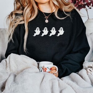 Op de afbeelding: Zwart sweatshirt met vier witte spookmotieven over de borst. De geesten zijn afgebeeld in een eenvoudige cartoonstijl. Het sweatshirt wordt gedragen en de achtergrond omvat een zachte deken en een mok met bloemenpatroon.
