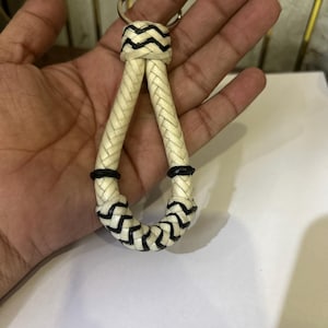 Puede incluir: Un llavero en forma de lágrima que presenta un cordón trenzado de color blanco roto con detalles en negro. El diseño incluye una anilla metálica circular en la parte superior, con detalles trenzados negros en la unión y en la curva inferior.