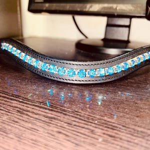 May include: Black leather browband with a curved design, decorated with a row of light blue and clear crystals. The crystals are set in a silver-coloured metal. Designed for equestrian use.